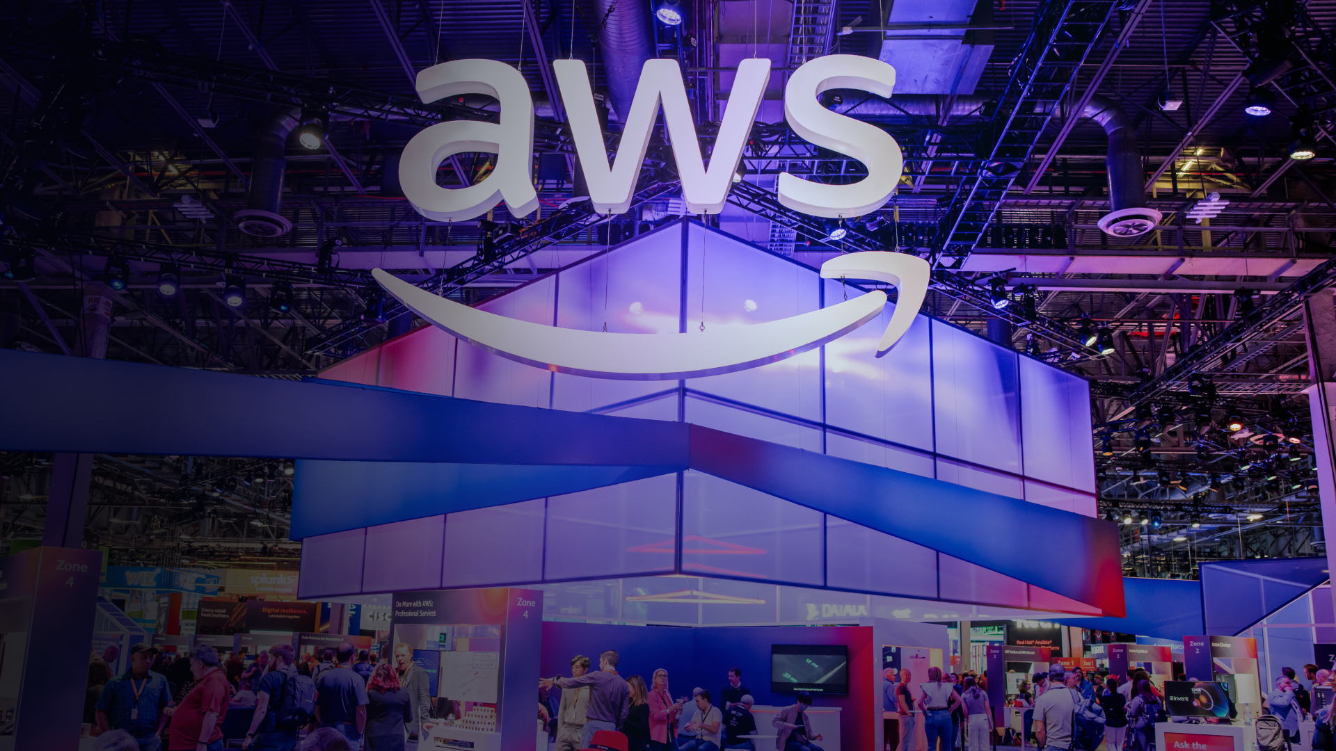 AWS booth at a conference or trade show featuring the iconic AWS logo and smile design suspended above a multi-level exhibition space with purple and blue gradient lighting, surrounded by attendees exploring various demo stations.